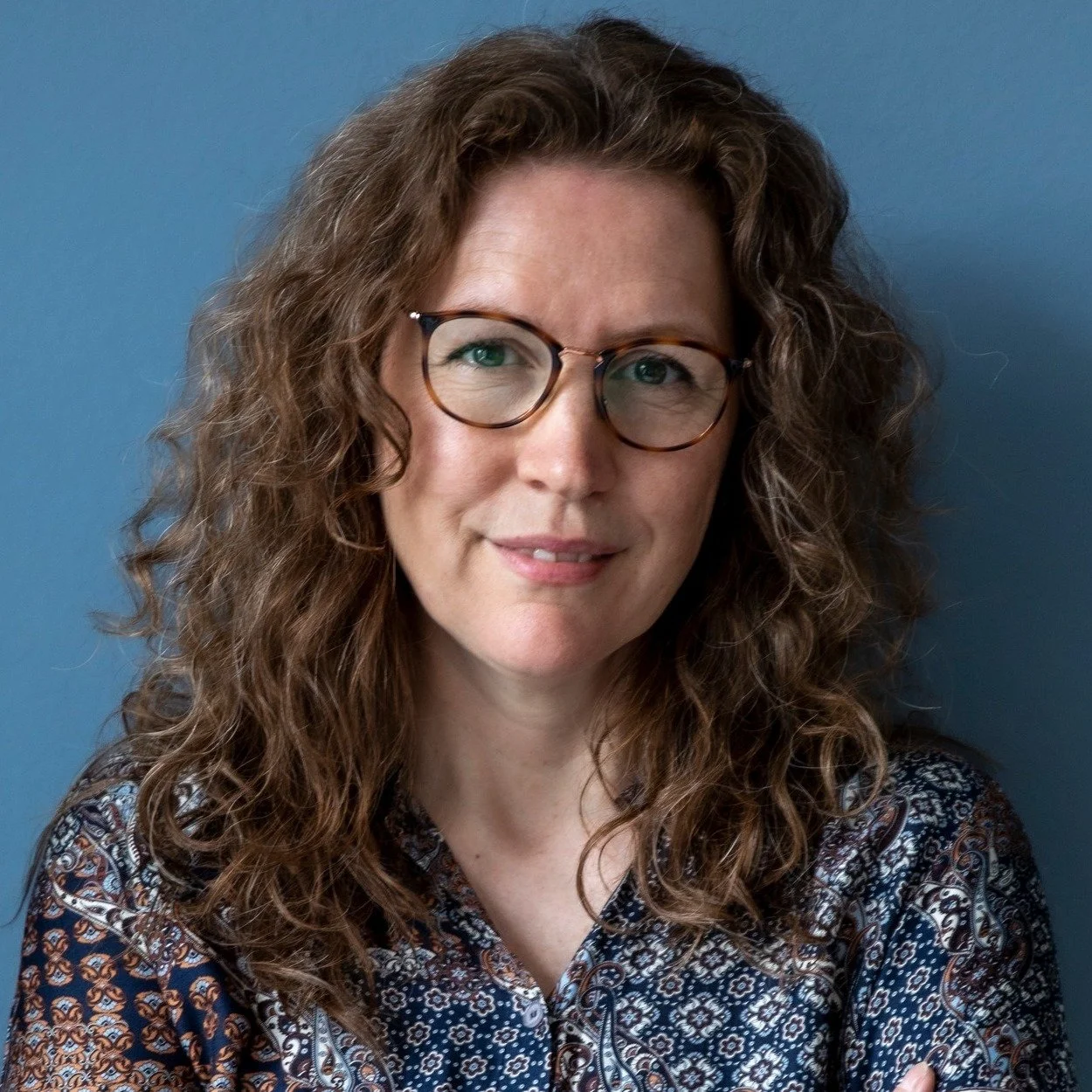 Portrætfoto af en kvinde med brune, krøllede hår, ser venlig ud, bærer briller og en blomstret bluse, står foran en blå baggrund.