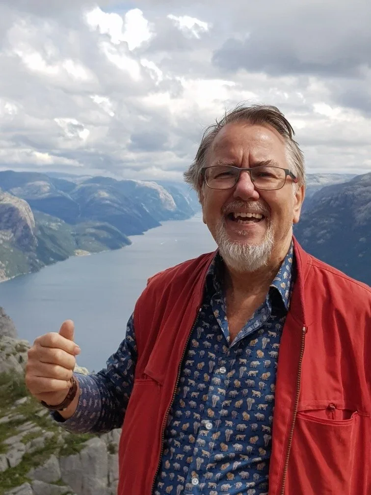 En glad mand med gråt skæg, briller og en rød vest står foran en fjord med bjergskaldoer under en overskyet himmel.
