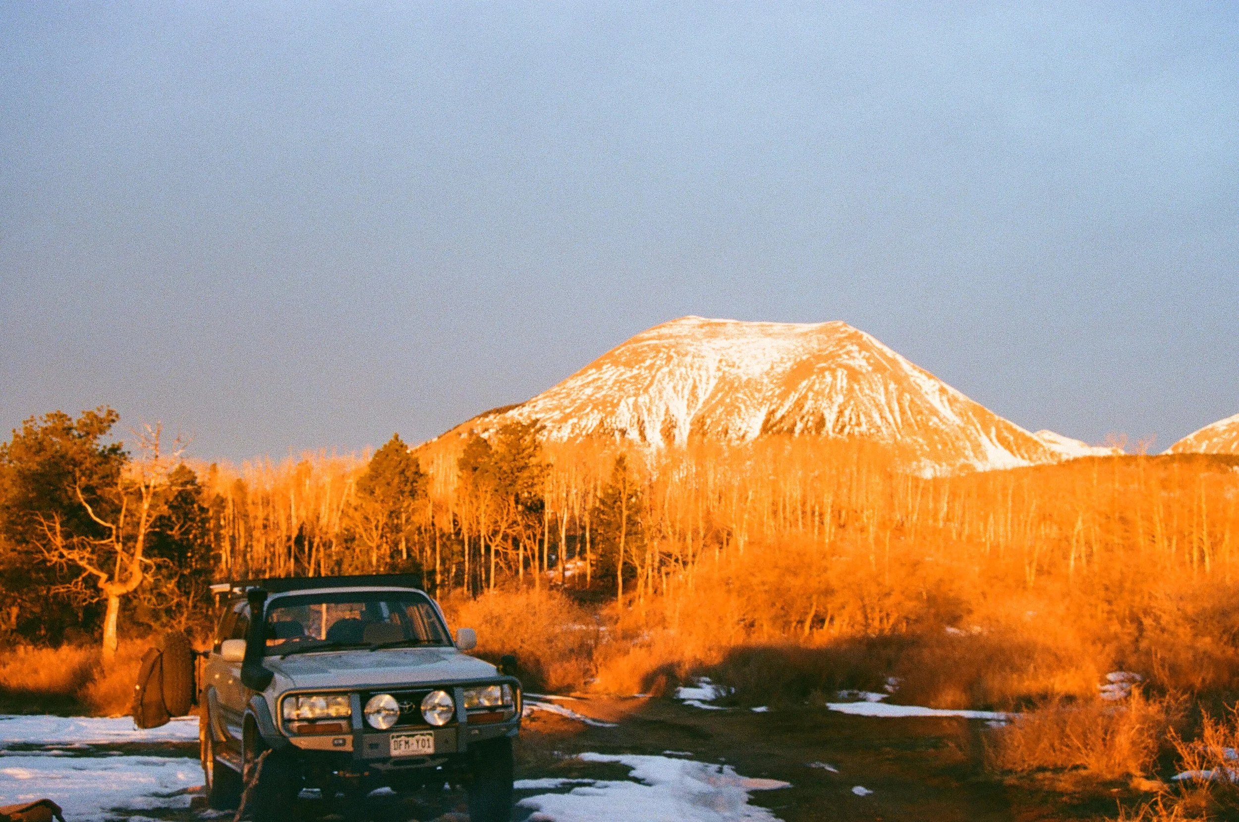 A vintage SUV parked on a snowy dirt road with a snow-capped mountain in the background during sunset.