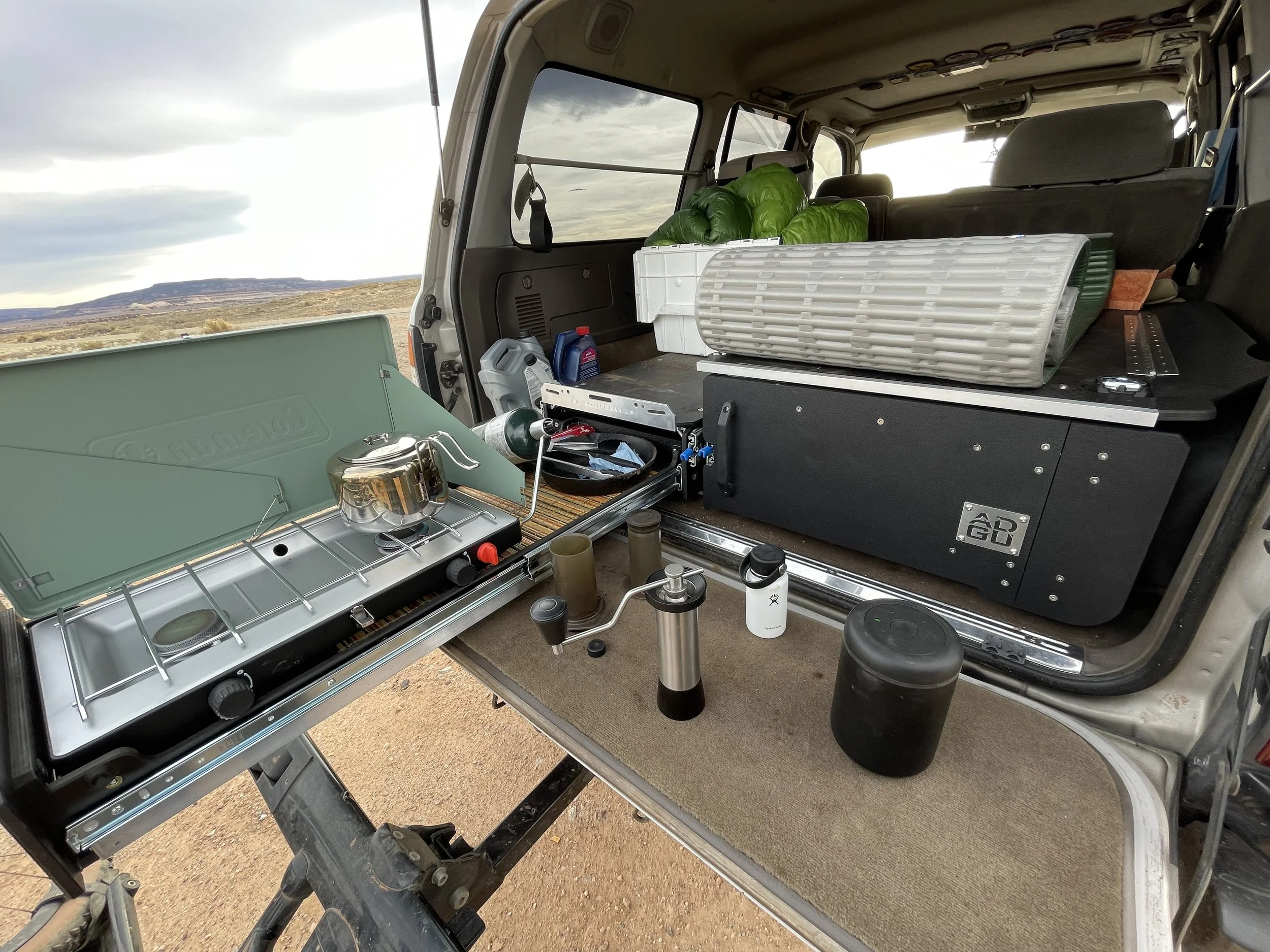 Rear interior of an SUV converted into a camping or outdoor cooking setup, featuring a portable stove, insulated food containers, water bottles, and greenery outside.