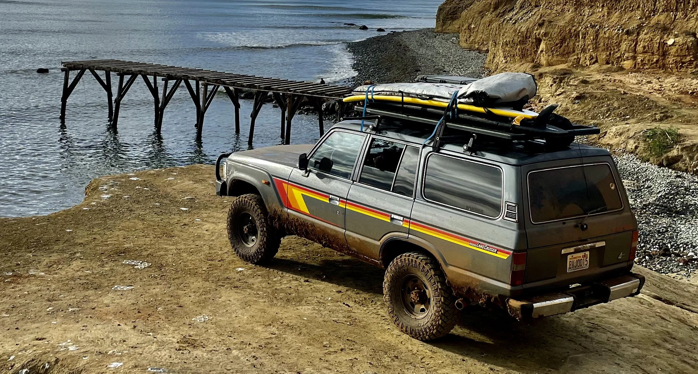 A silver off-road vehicle with roof-mounted gear and a surfboard, parked on a dirt patch near a rocky shoreline, with a wooden pier extending into the water in the background.