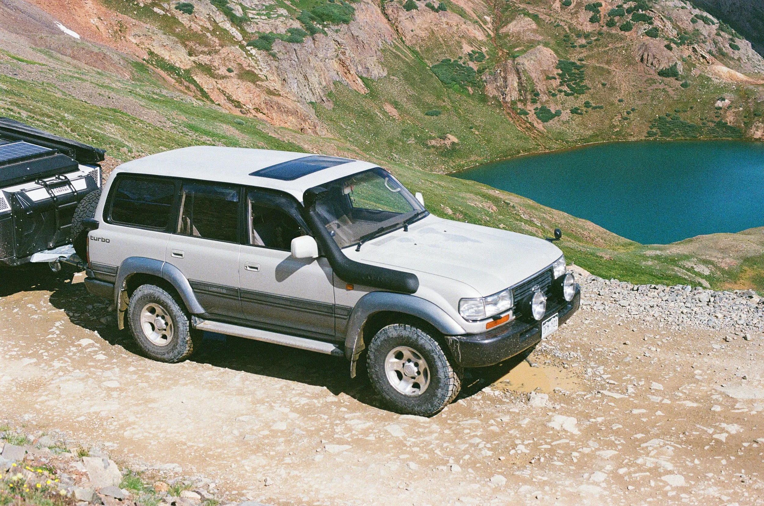 A white SUV parked on a dirt trail near a mountain lake with rocky and green slopes.