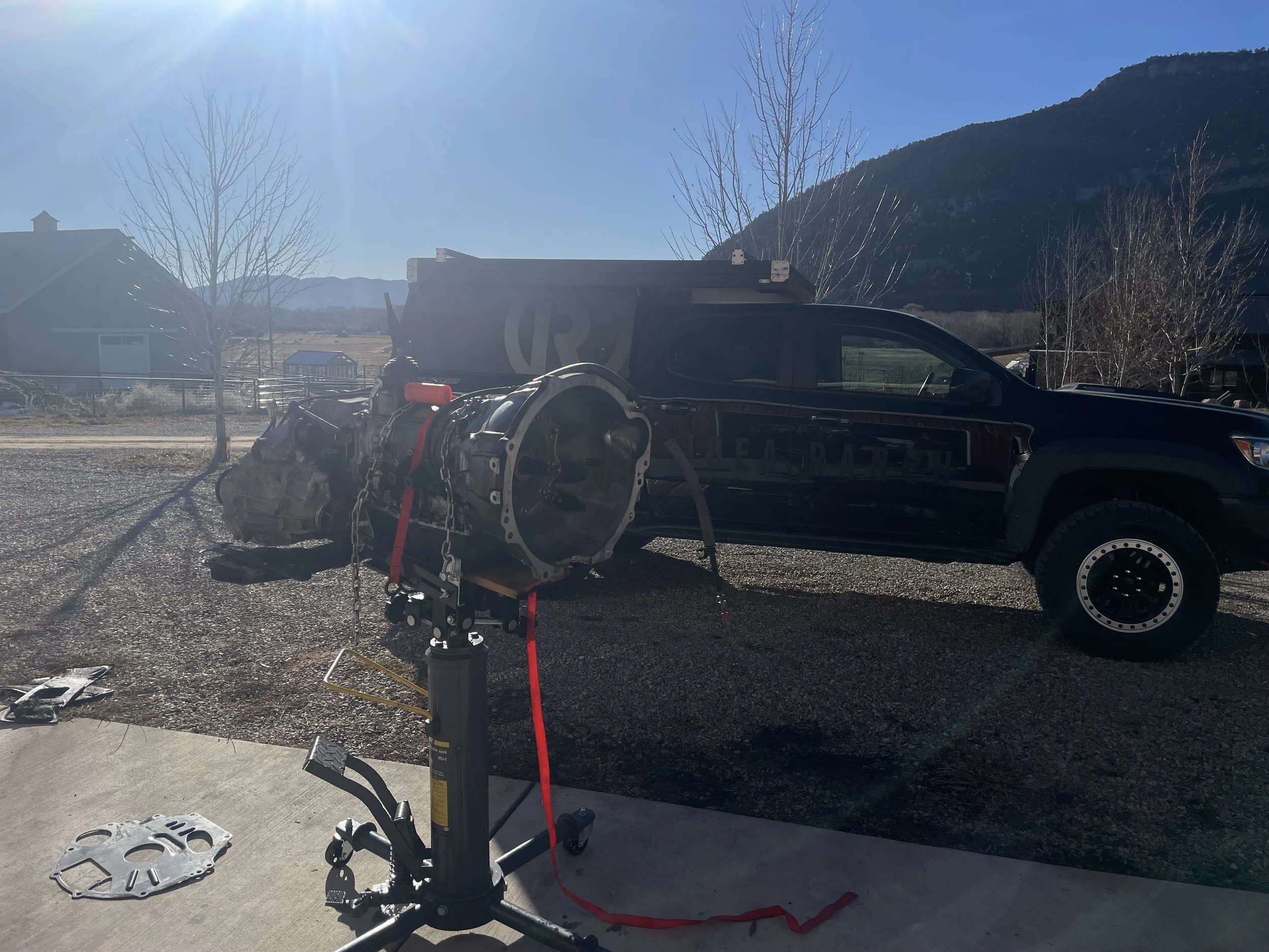 A black vehicle with a large logo parked outside on a dirt lot during daytime, with engine or transmission being worked on a stand in front of it, and mountains visible in the background.