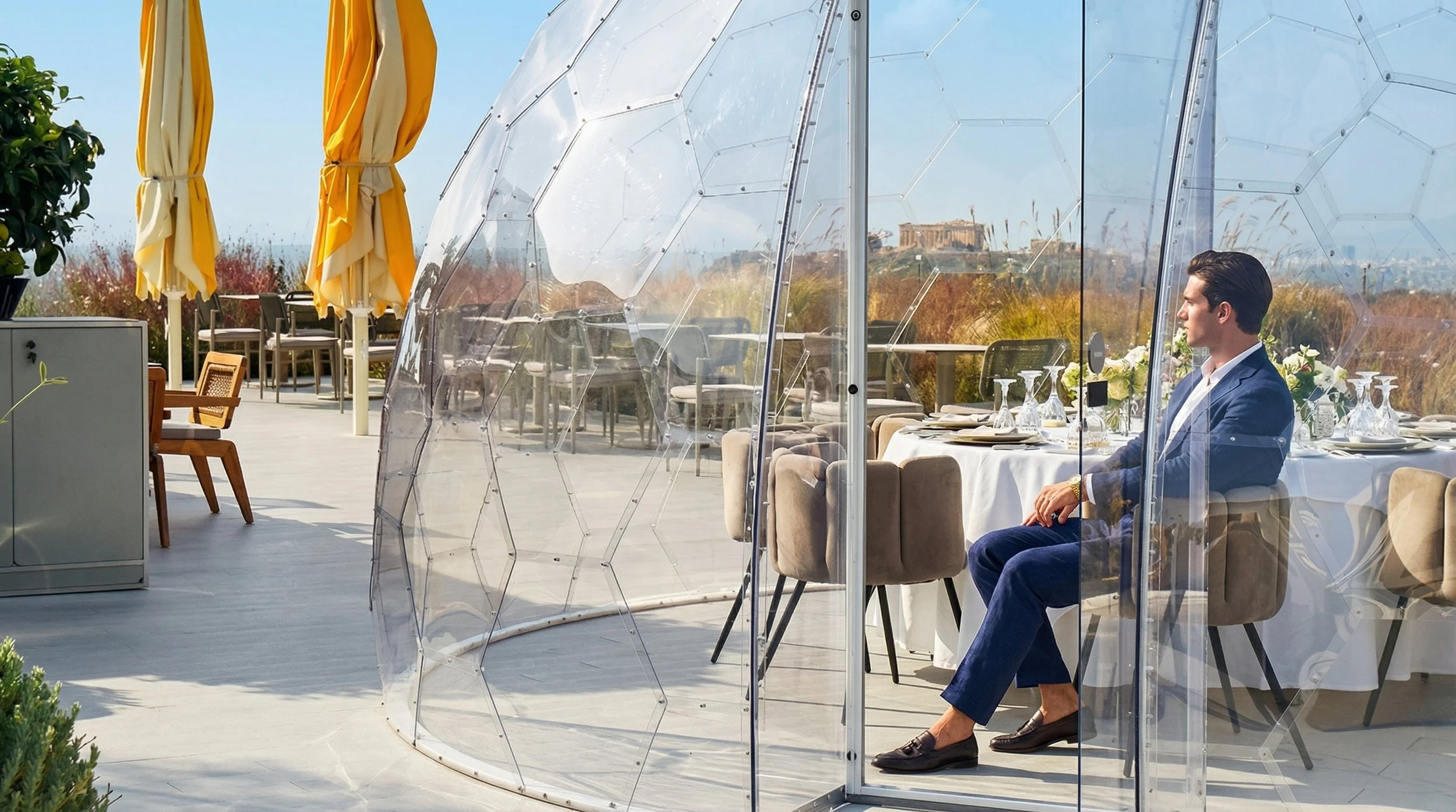 A man enjoying a romantic dinner inside a transparent heated dome on the rooftop of St. George Lycabettus Hotel with a daytime view of the stunning  Acropolis.
