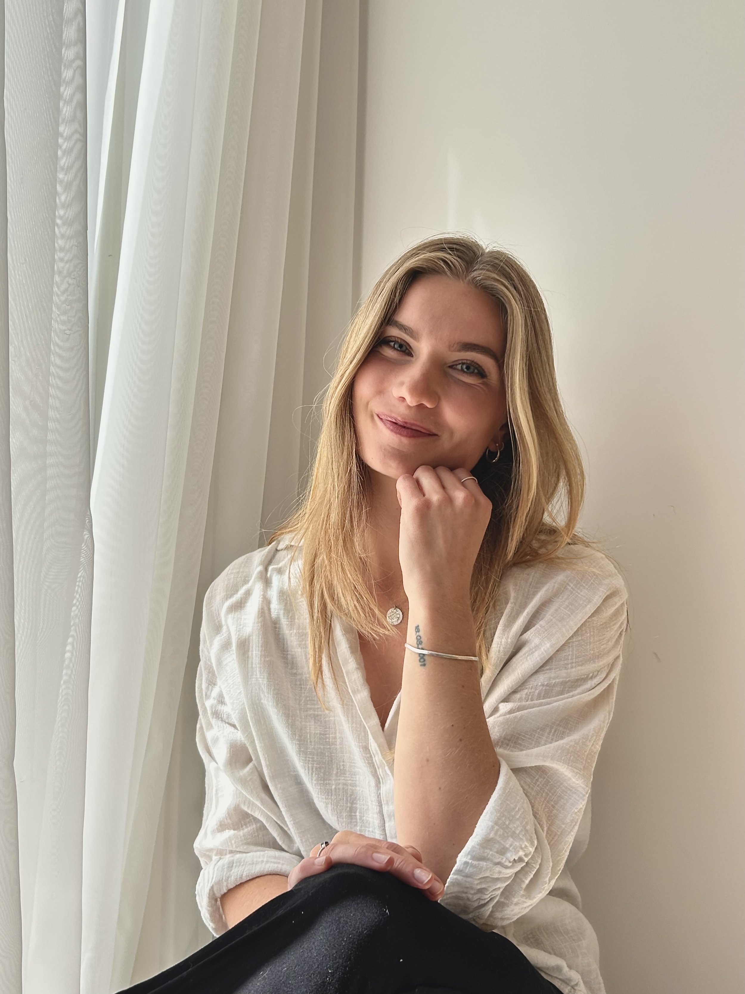 A young woman with long blonde hair, wearing a white shirt, smiling gently, resting her chin on her hand near her face, sitting near white curtains and a plain white wall.