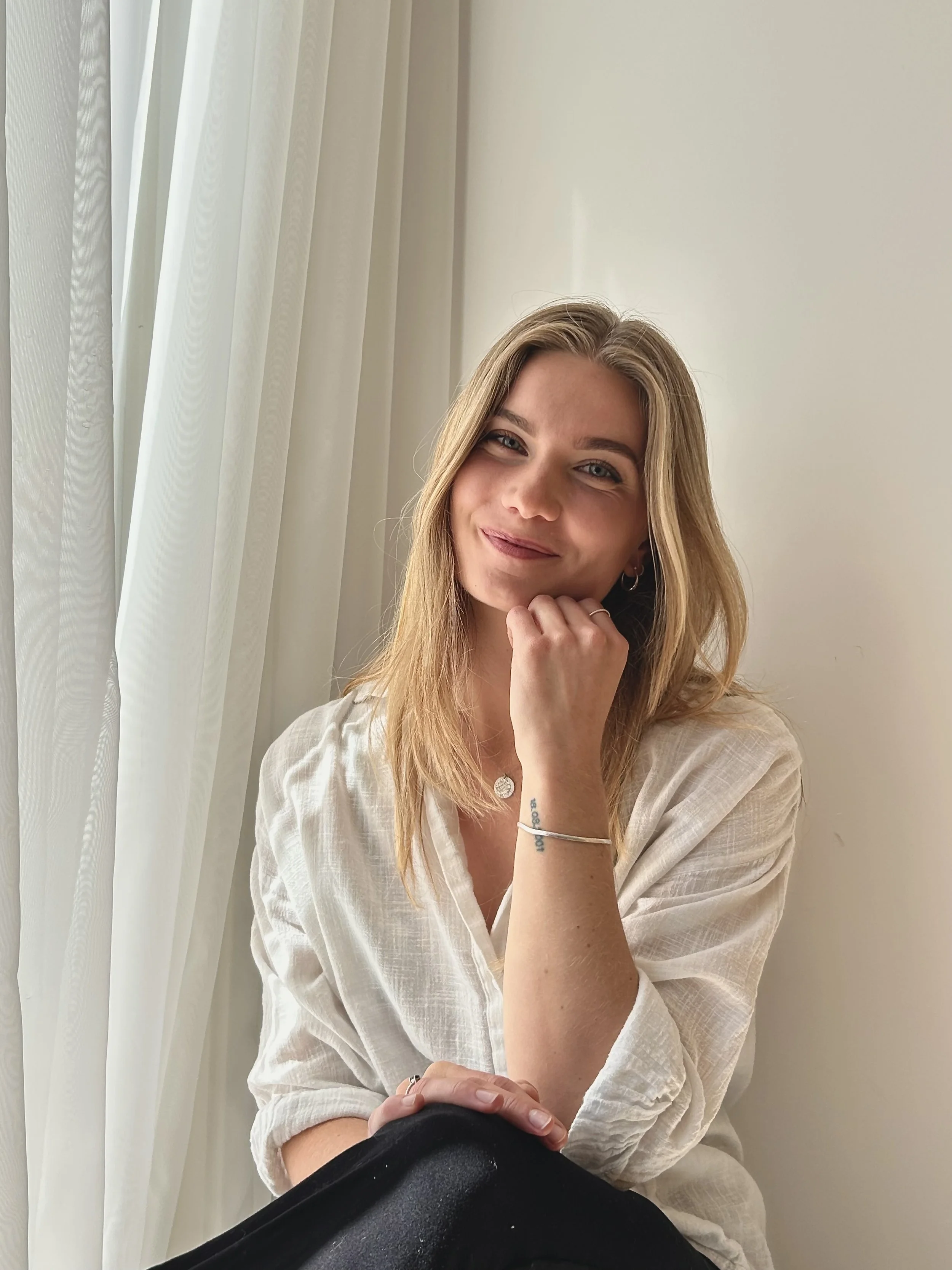 A young woman with long blonde hair, smiling softly, resting her face on her hand, sitting near white curtains and a plain white wall, wearing a light-colored linen shirt and jewelry.