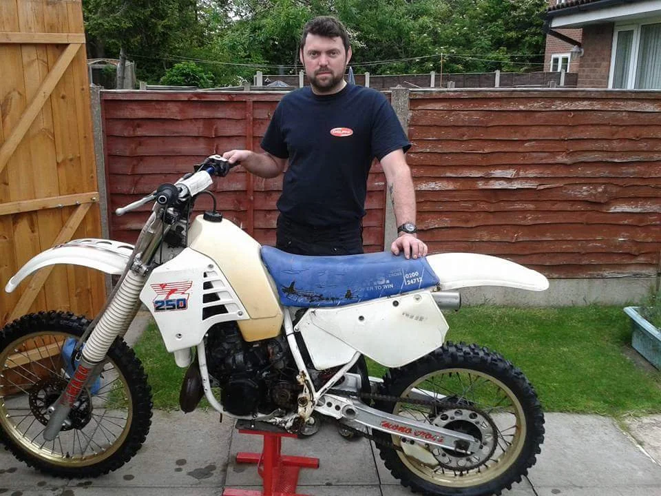 A man with dark hair and beard wearing a black t-shirt stands outdoors next to a vintage off-road motorcycle on a red stand. The motorcycle is white with some black parts and has a blue seat. Behind is a wooden fence and some greenery.