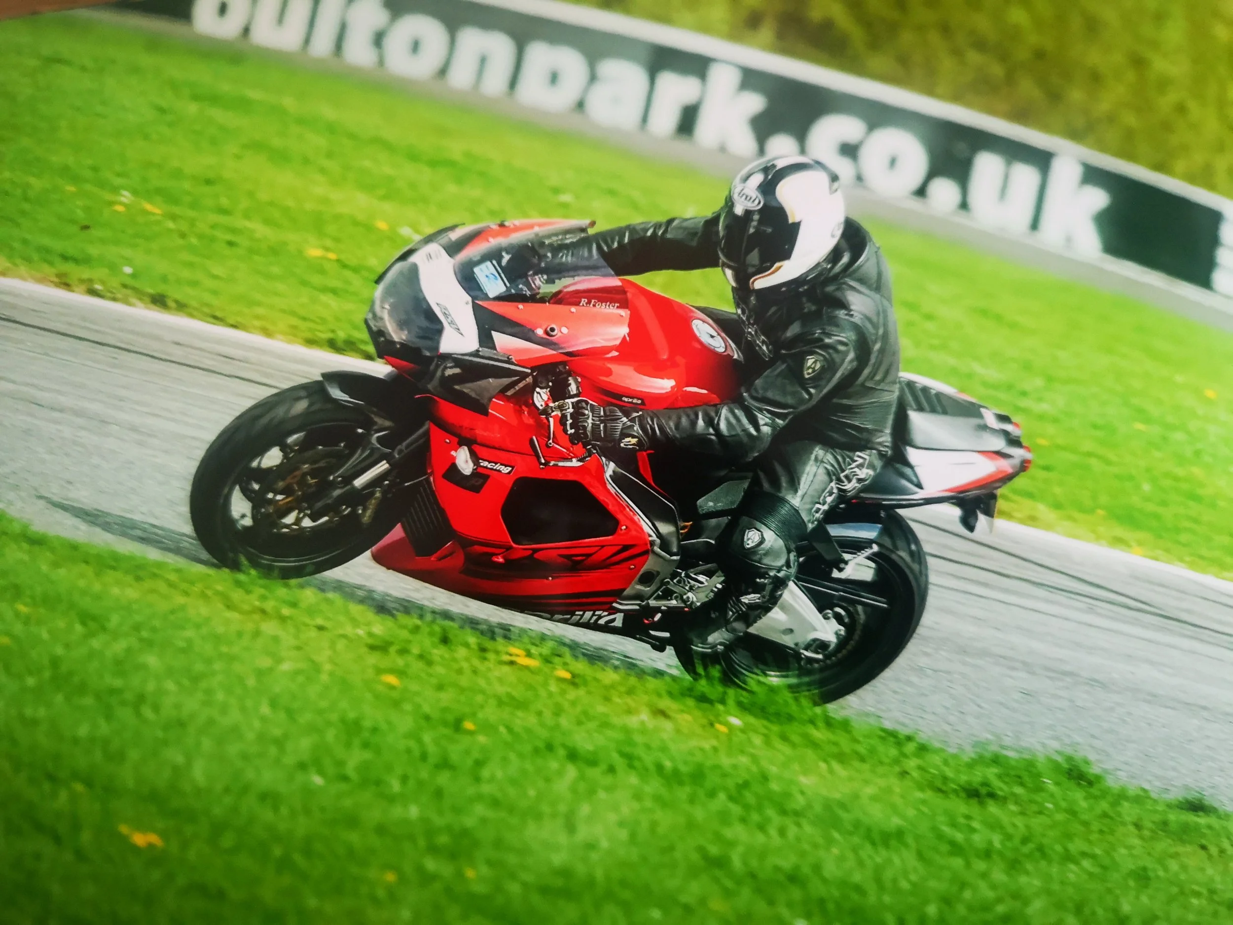 A motorcyclist wearing black riding gear and a helmet leaning into a turn on a red and black sportbike on a race track, with green grass and a banner in the background.