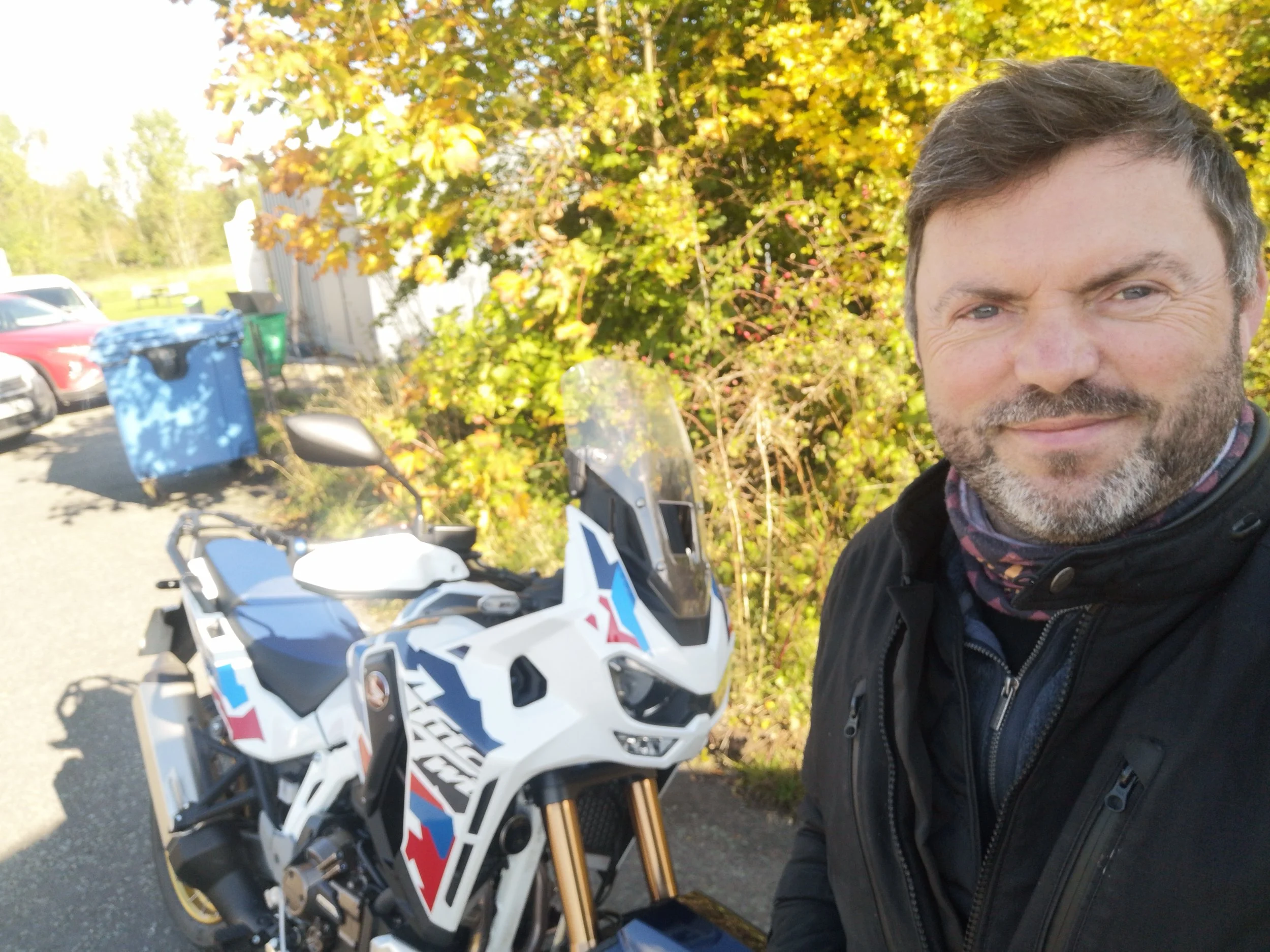 A man taking a selfie outdoors next to a white police motorcycle with a clear windshield, with trees and cars in the background, during daytime.