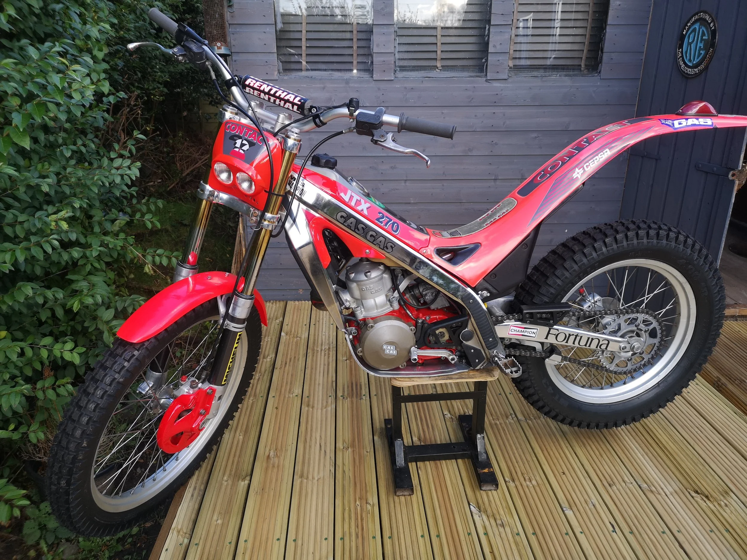 A red and silver motocross bike with large tires, positioned on a black stand on a wooden deck, with a gray wooden building and green shrubbery in the background.