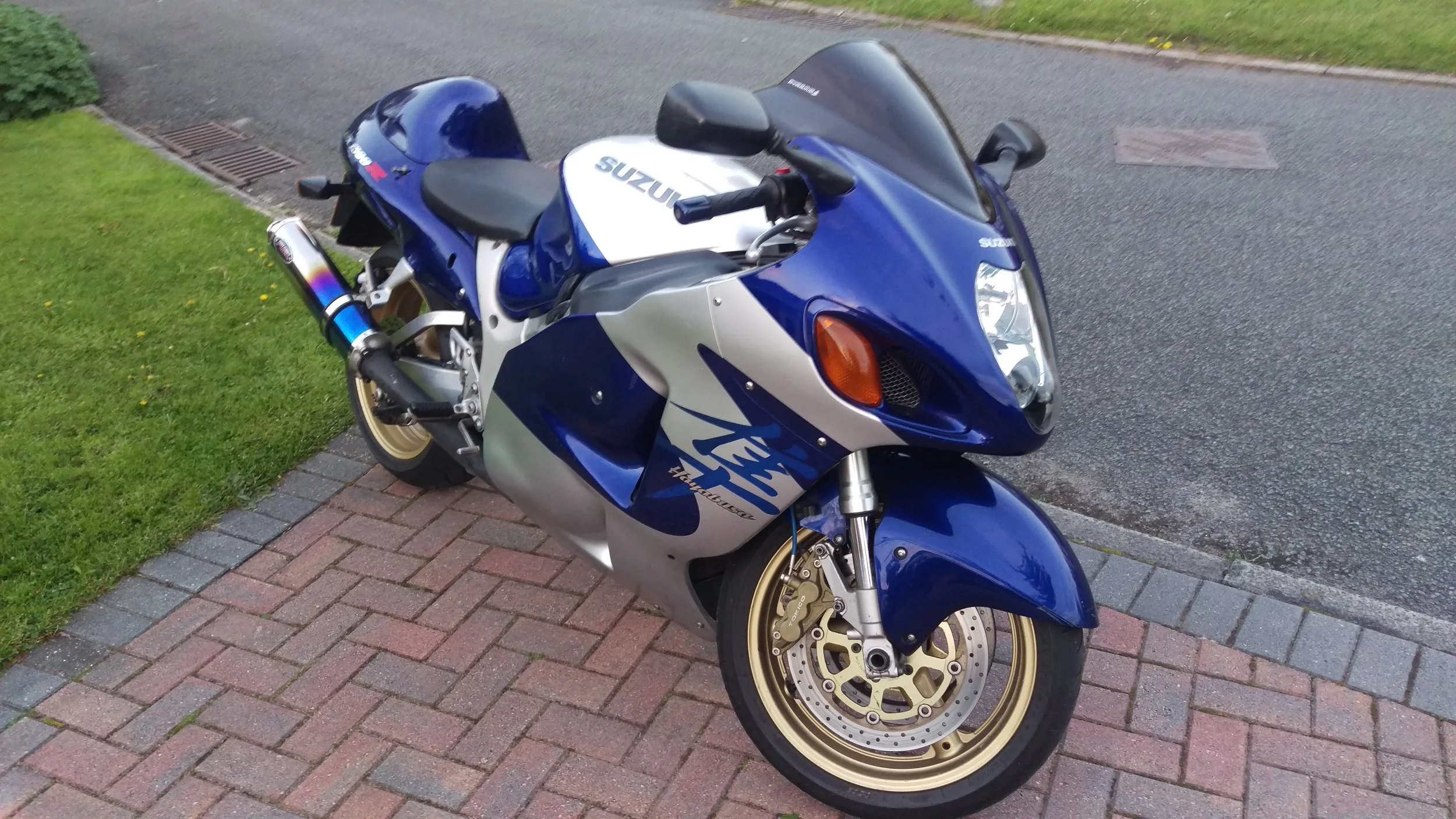 A blue and white Suzuki Hayabusa sport motorcycle parked on a brick driveway next to a grassy lawn.