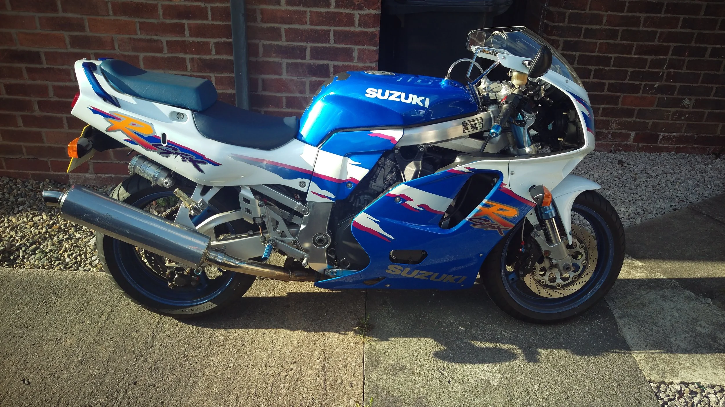 A blue and white Suzuki sport motorcycle parked on a concrete driveway with a brick wall in the background.