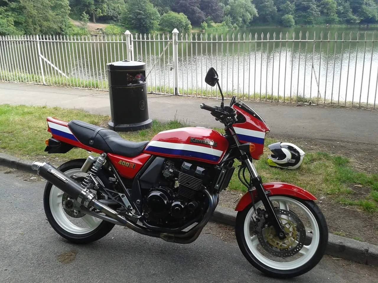 Red Kawasaki motorcycle with white and blue accents parked by a sidewalk next to a lake, with a white helmet on the ground nearby and a black trash can in the background.