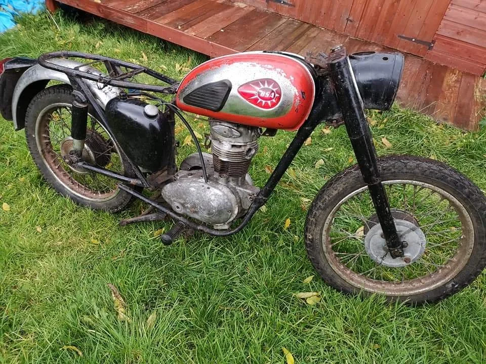 An old, vintage motorcycle with a red and silver gas tank, black frame, and black front forks, parked on green grass next to a wooden fence.