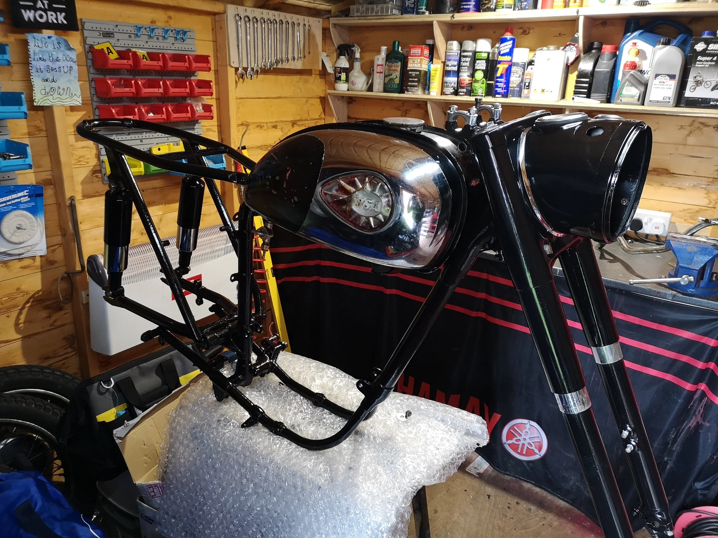 A motorcycle frame and fuel tank being assembled in a workshop, with tools and supplies on the shelves behind.
