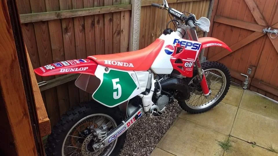 Red and white dirt bike with racing stickers and a green number 5 plate, parked in a backyard.