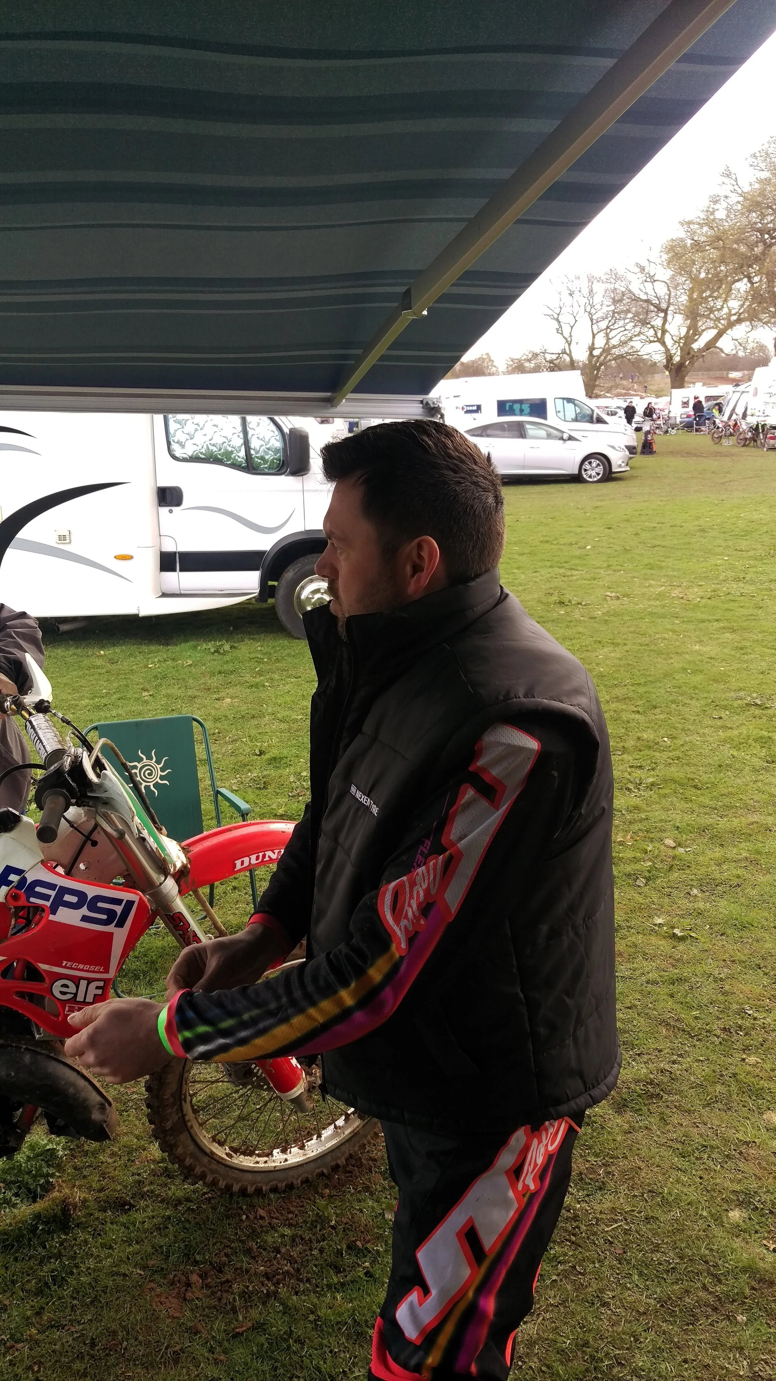 A man in racing gear standing next to a dirt bike under a canopy at an outdoor event, with RVs and trailers in the background.