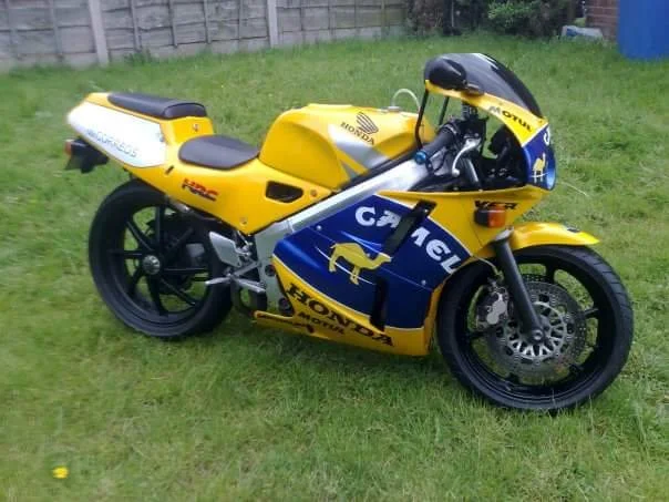 Yellow and blue racing motorcycle with Honda branding, parked on grass with a fence in the background.