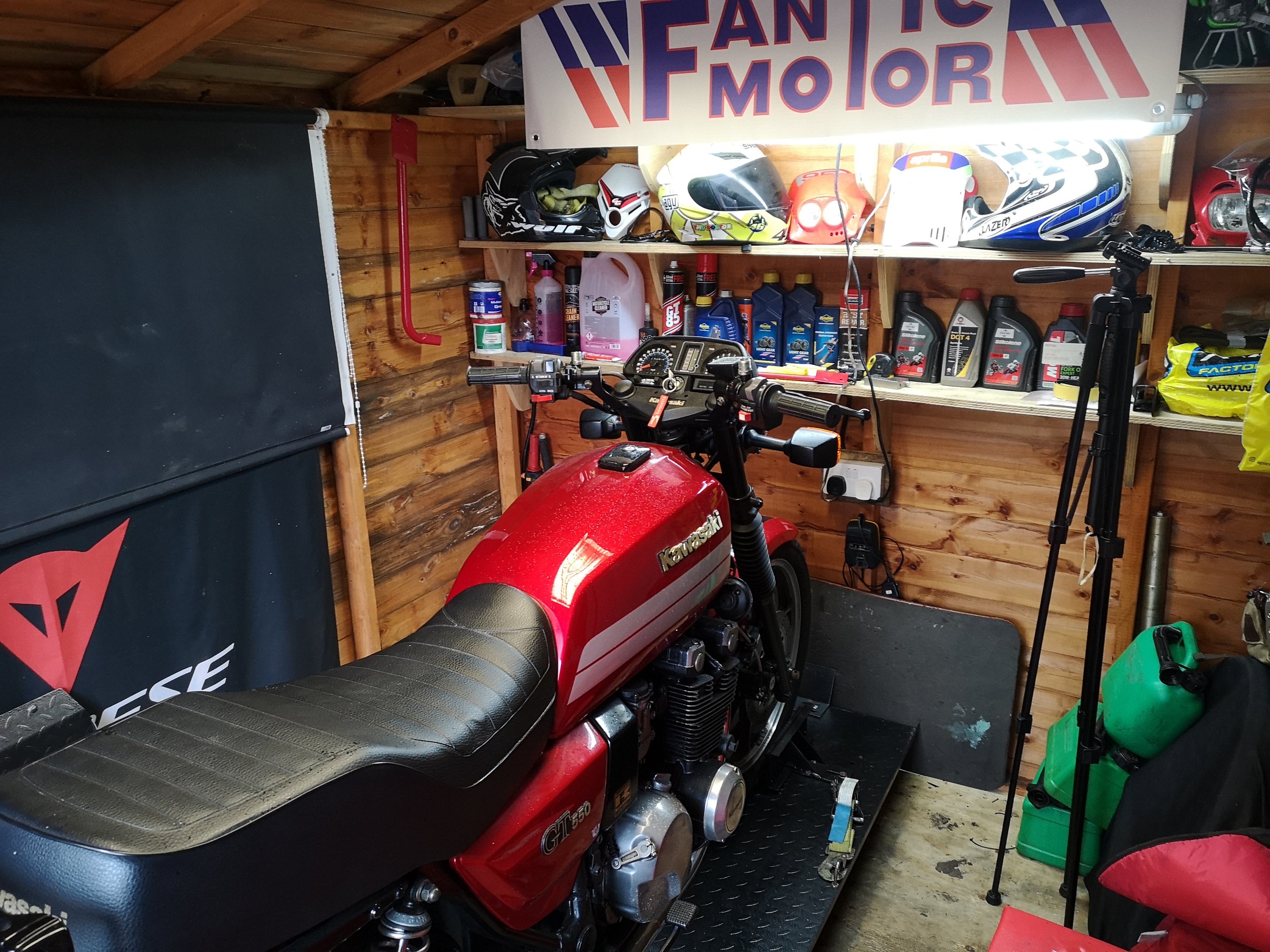 A red Kawasaki motorcycle parked inside a wooden garage. The garage has a shelf holding various helmets, motor oil, and other supplies. There is a Dainese banner hanging on the wall.