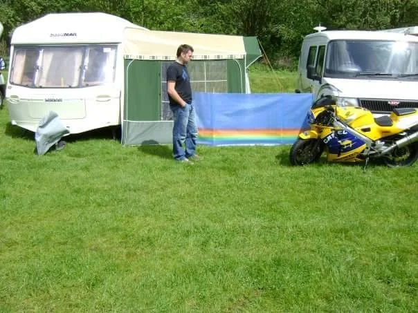 A man standing on grass near a white camper trailer and a white van, with a colorful rainbow flag hanging between the two vehicles. There is a yellow and blue motorcycle parked beside the van in a grassy outdoor setting.