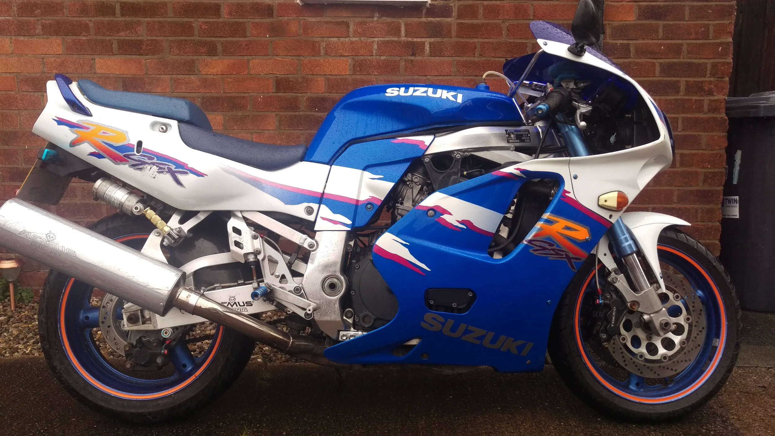 Blue and white Suzuki sport motorcycle with orange accents parked against a brick wall on a wet ground.