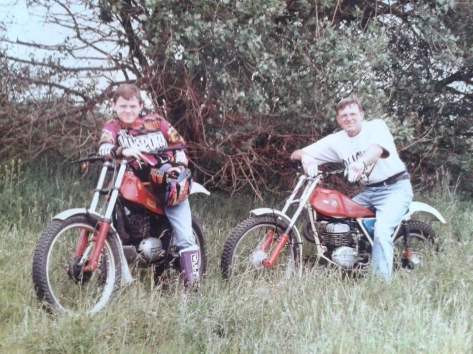 Two children riding vintage red motorcycles outdoors on grass with trees and bushes in the background.