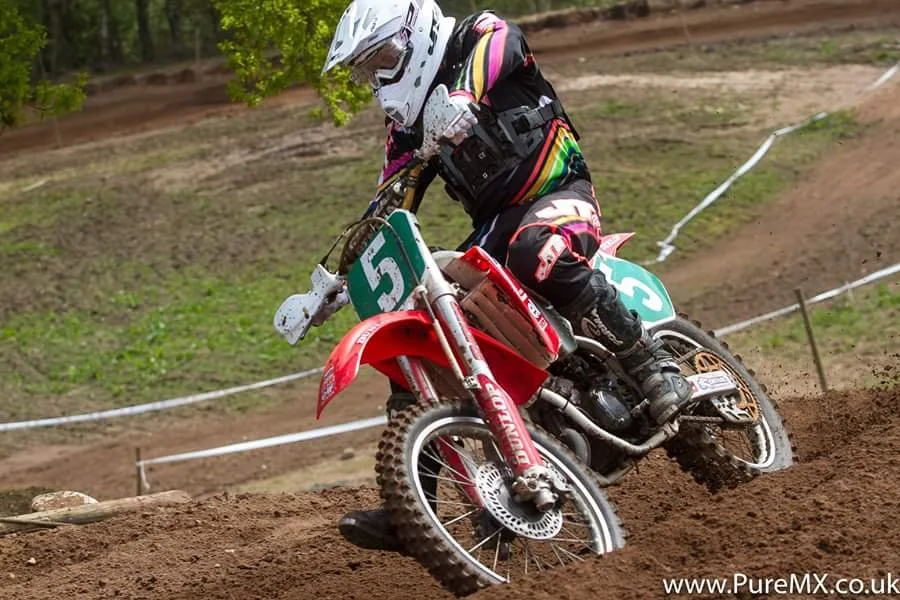 A motocross rider in full gear leaning into a turn on a dirt track, riding a red and white dirt bike with green and blue number plates.