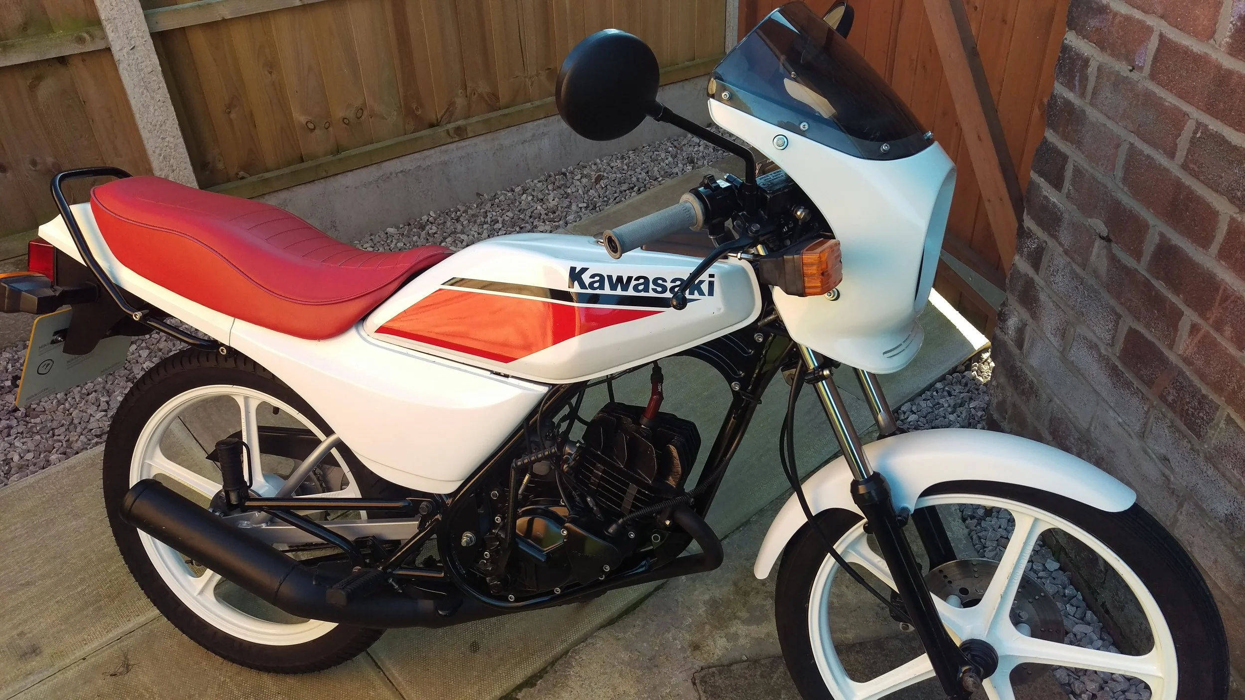 A vintage Kawasaki motorcycle with a red seat and white body, parked on a concrete surface next to a wooden fence and brick wall.