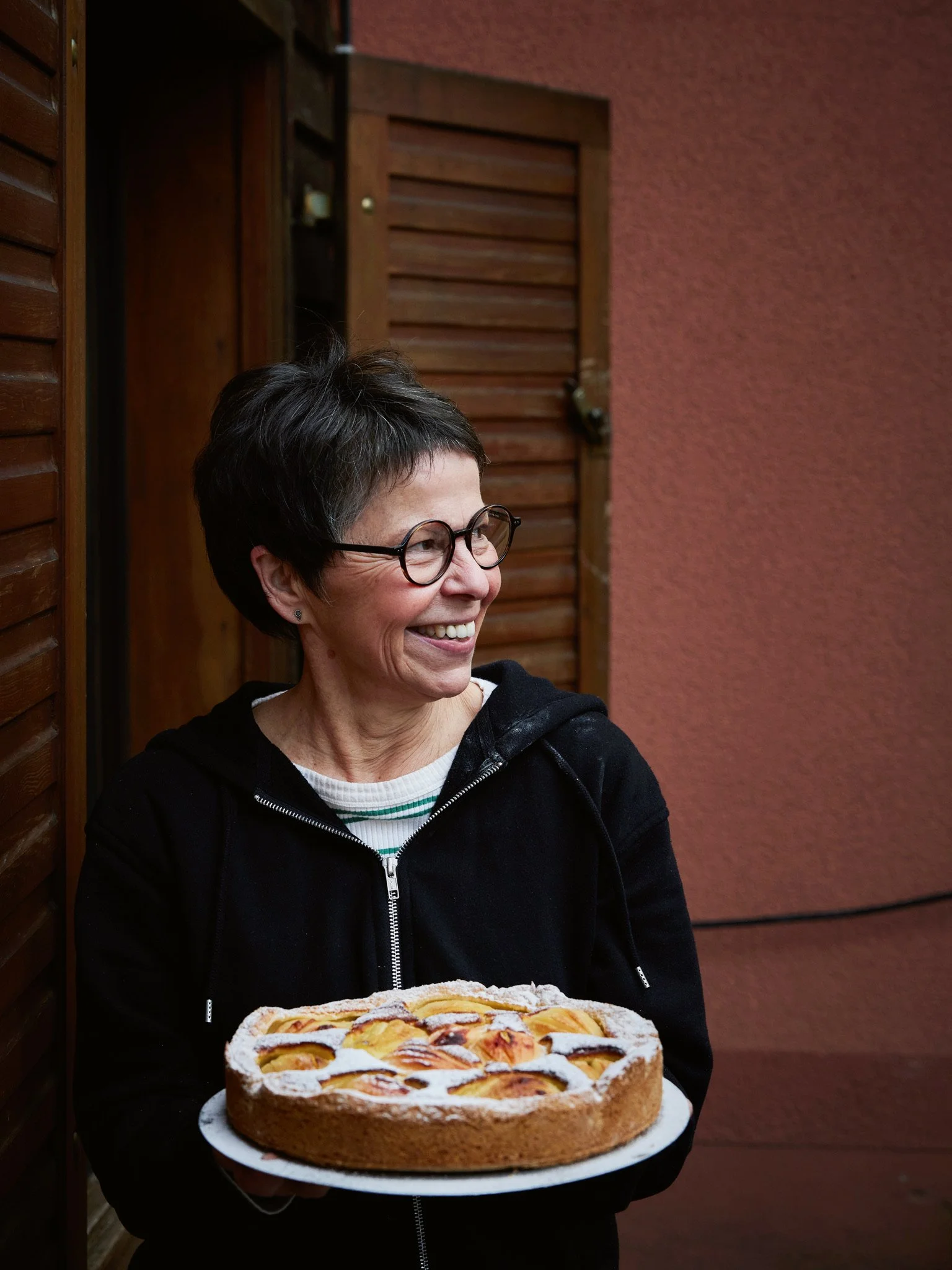 Eine lachende Frau mit Brille hält einen Kuchen mit Obst und Puderzucker vor einem roten Wand und Holzfensterläden in der Hand.