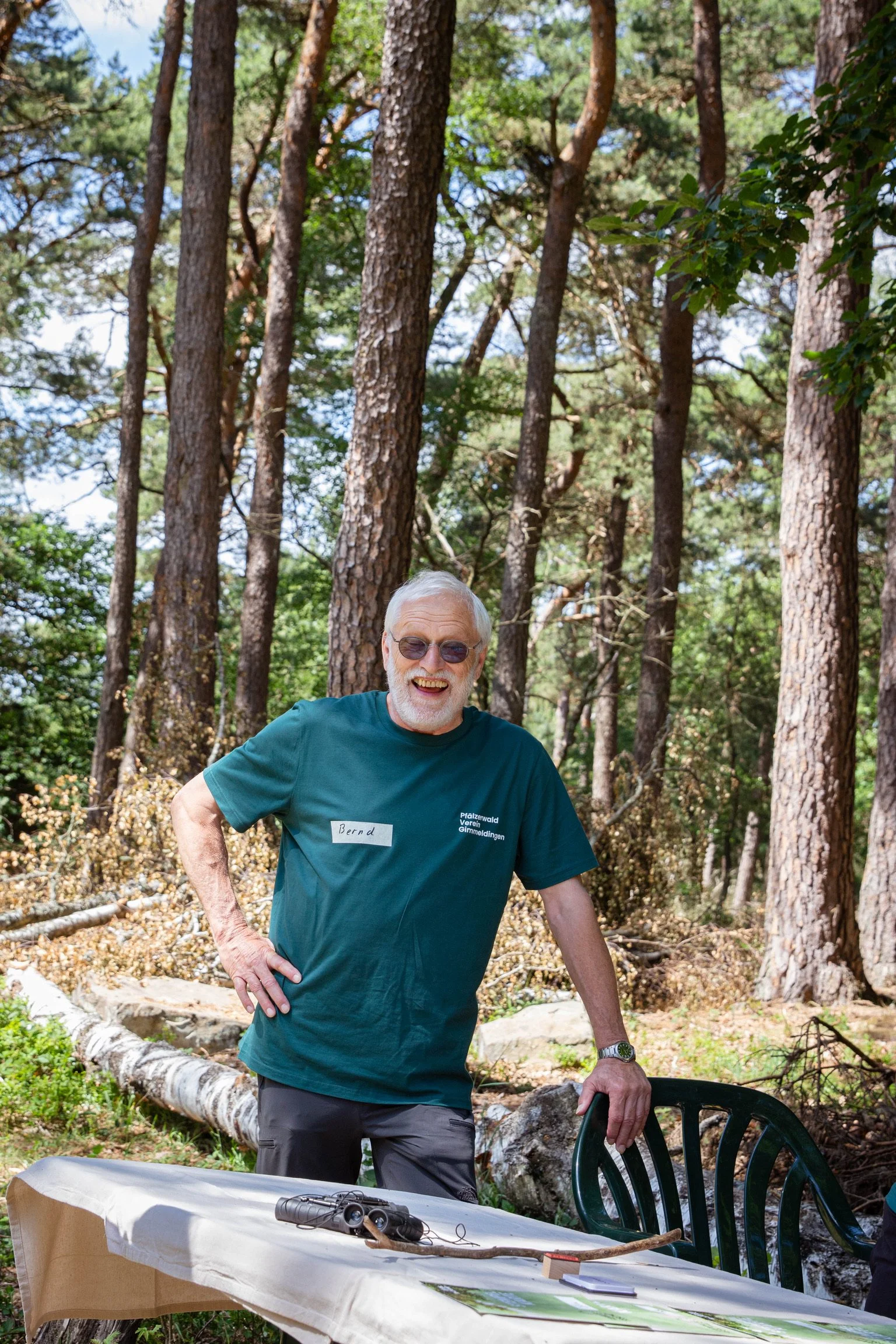 Älterer Mann mit Brille und Namensschild 'Bernd' steht im Wald vor einem Tisch mit einem Fernglas auf einer weißen Tischdecke, umgeben von Bäumen.