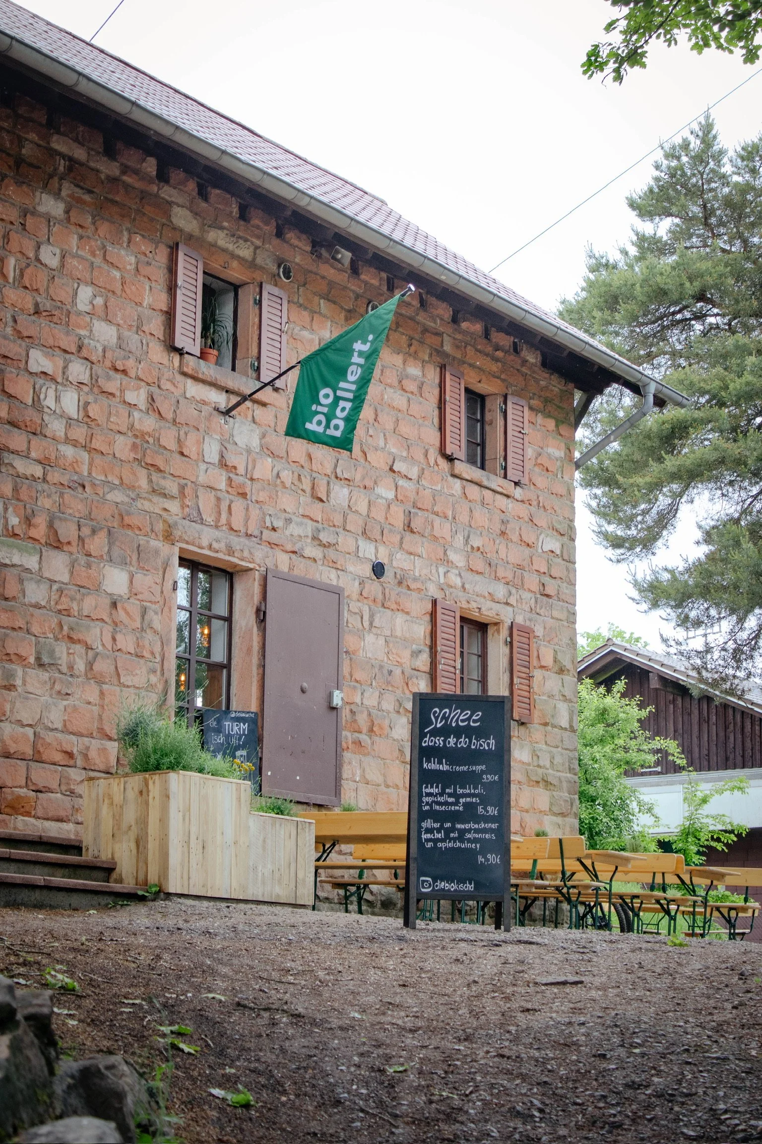 Ein Haus mit Steinmauer, offenen Fenster, grünem Banner mit weißer Aufschrift 'bio batt.' und einer Tafel mit Speisekarte draußen vor der Tür.