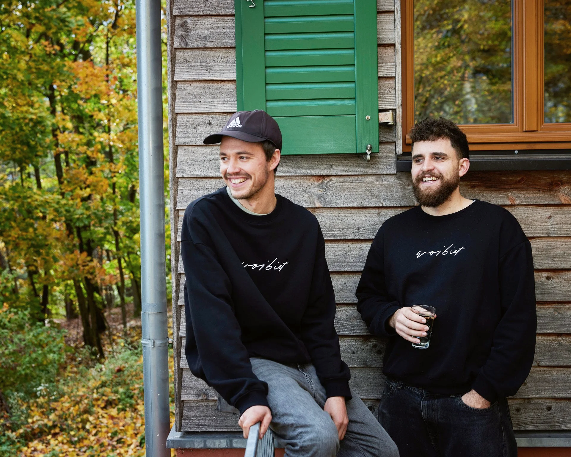 Sebi und Isaak, die in einer Herbstumgebung vorm Weinbiethaus stehen, tragen schwarze Sweatshirts mit weißen Schriftzug, einer hält ein Glas und der andere sitzt auf dem Geländer.