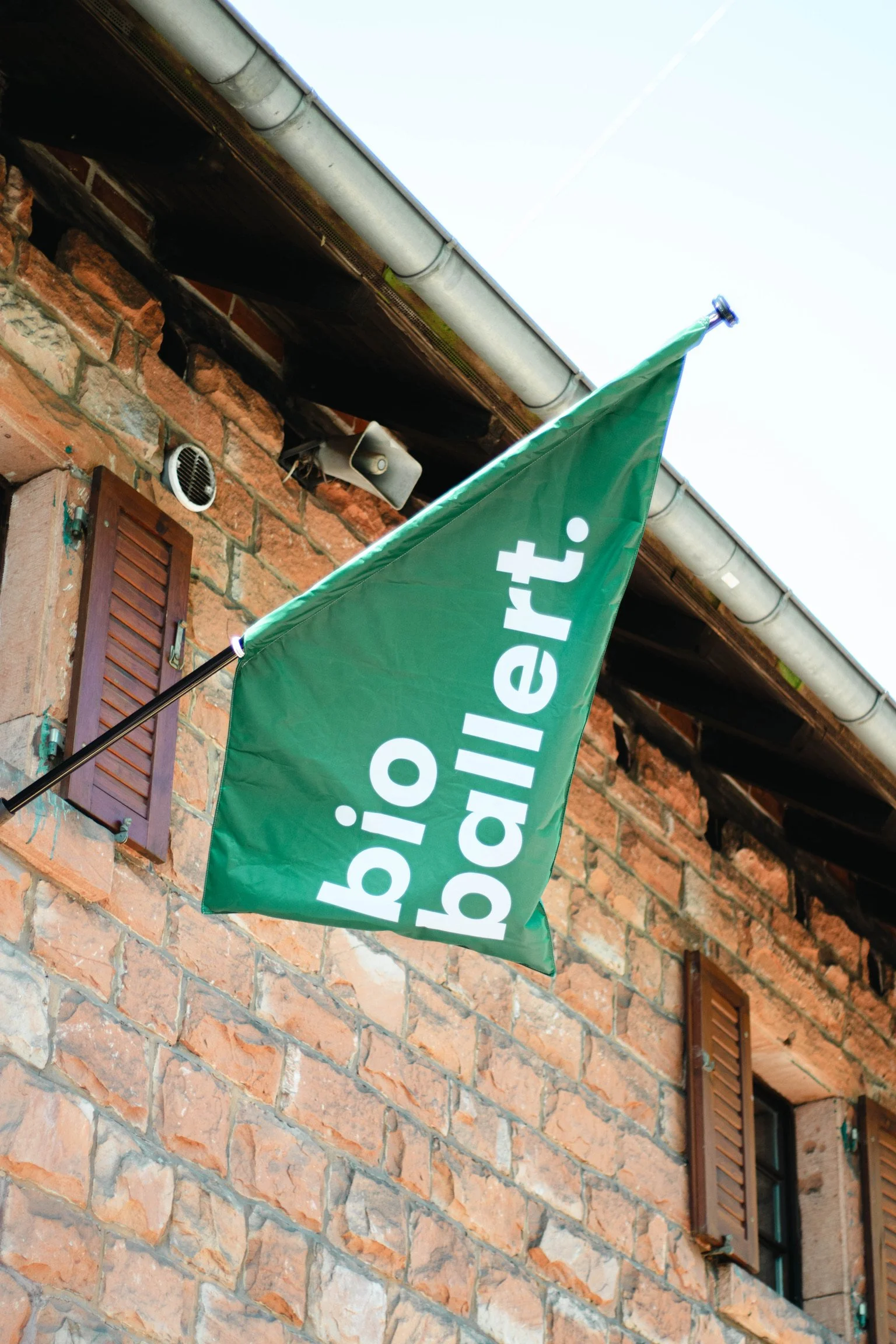 Grüner Flagge mit weißem Text 'bio ballert' hängt an einem Gebäude mit roten Ziegelwänden und Holzfensterläden.