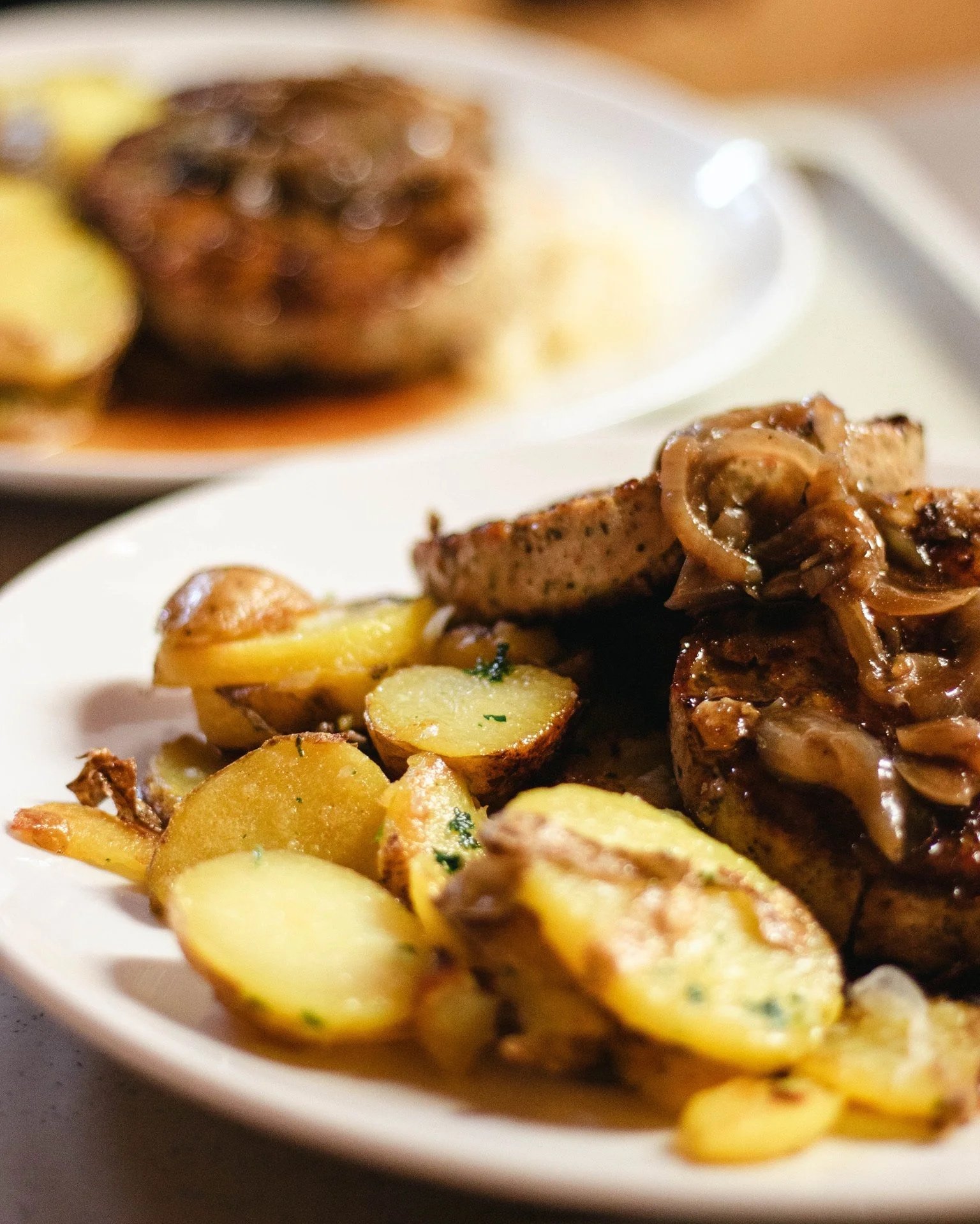 Hauptgericht mit Kartoffeln, gebratenem Fleisch und Zwiebeln, im Hintergrund weiteres Gericht mit Fleisch und Soße.