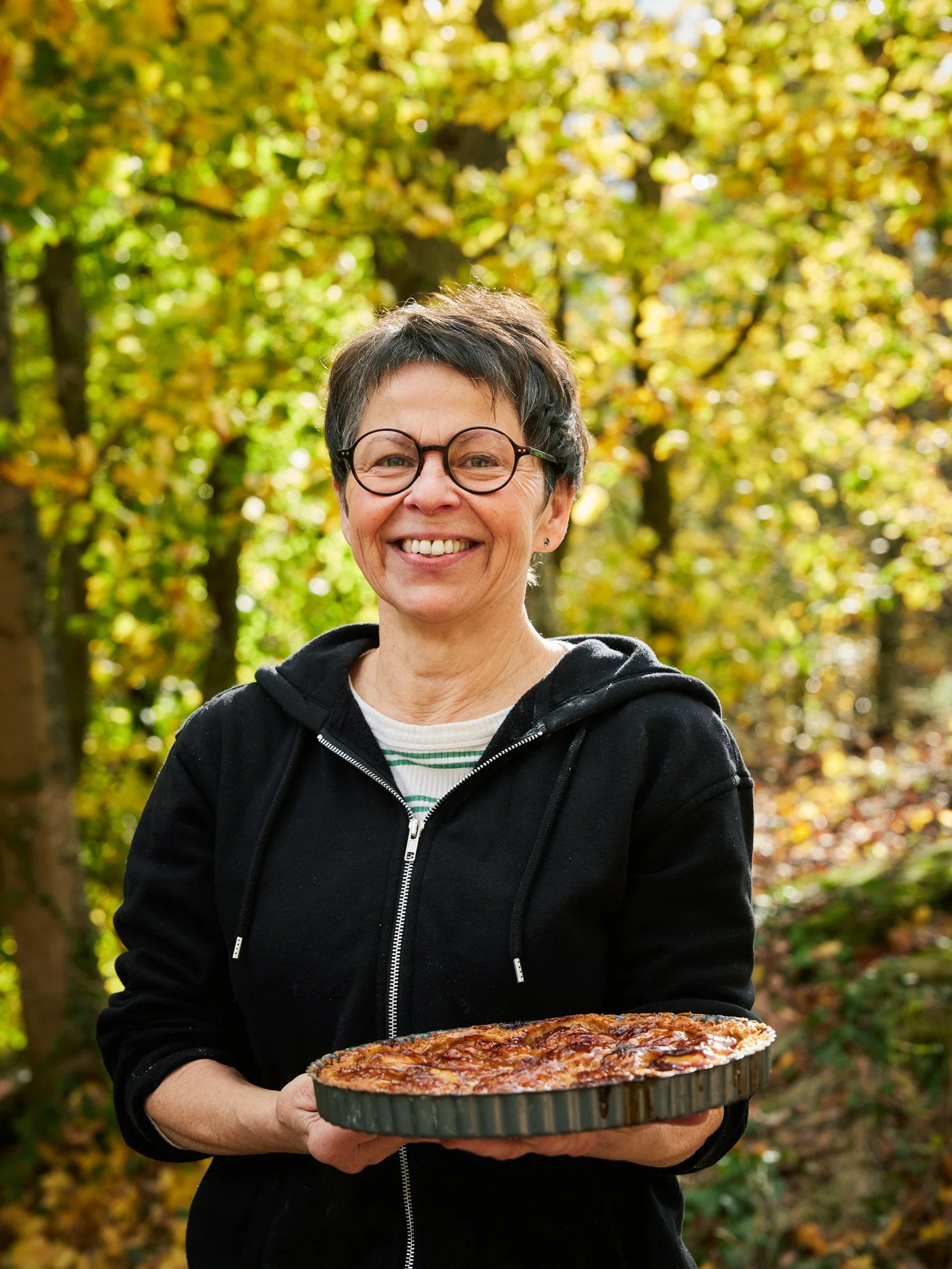 Eine lachende Frau trägt eine Tarte in einem schwarzen Tortenring in einem herbstlichen Wald.