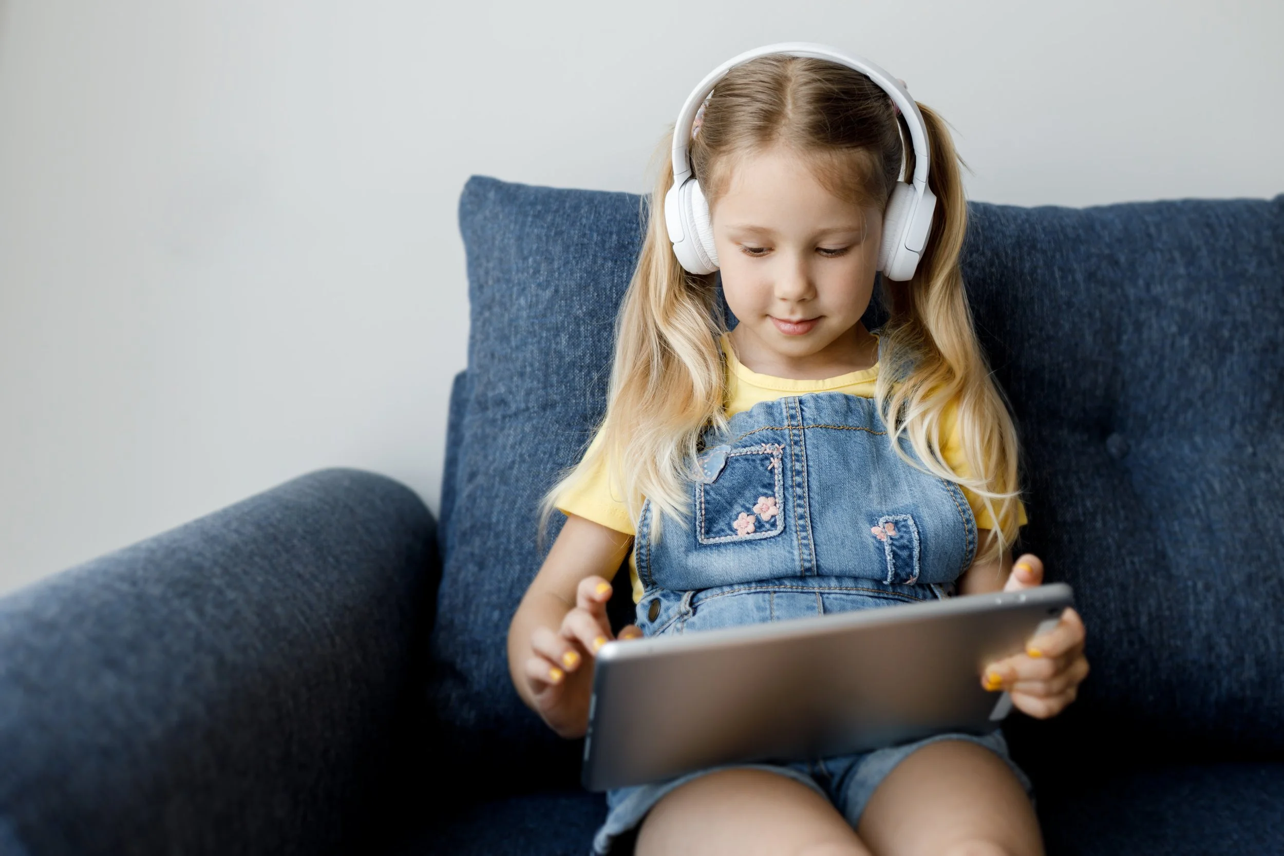 A young girl sitting on a blue couch, wearing white headphones, yellow shirt, and denim overalls, holding and looking at a tablet.