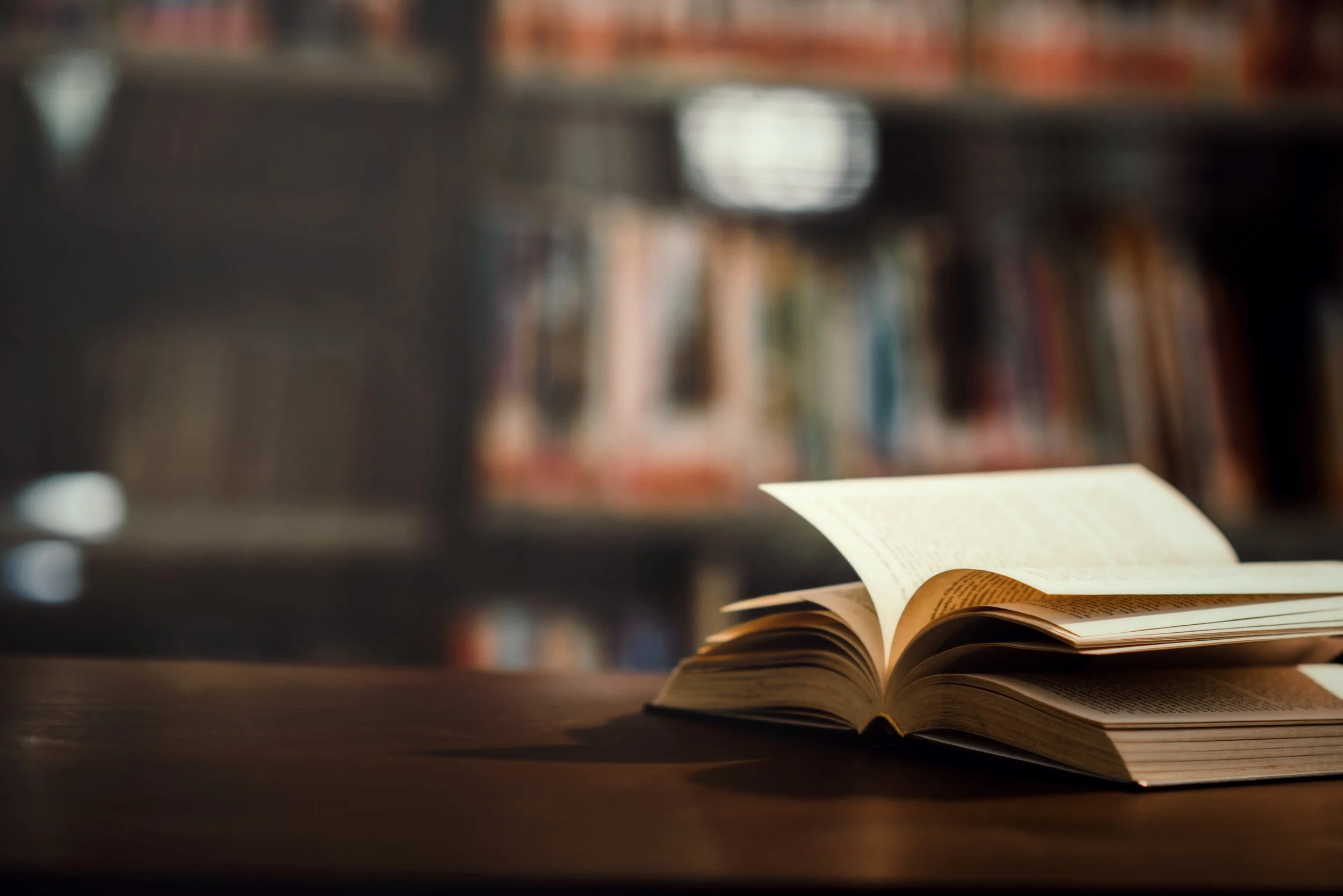 An open book on a wooden table in a library with bookshelves in the background.