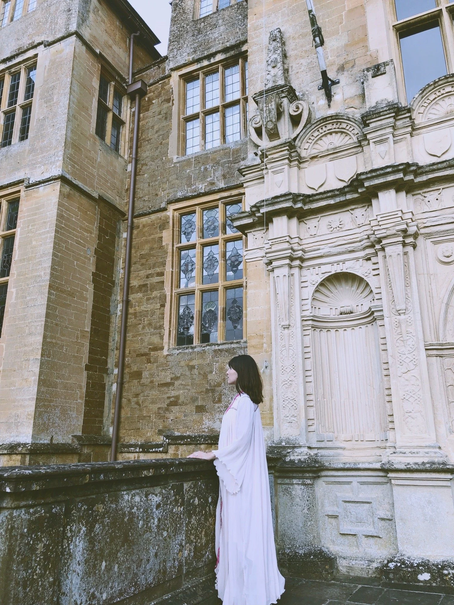 Woman in white dress standing on a stone balcony outside of an old historic building with stone walls, decorative stone architecture, and stained glass windows.
