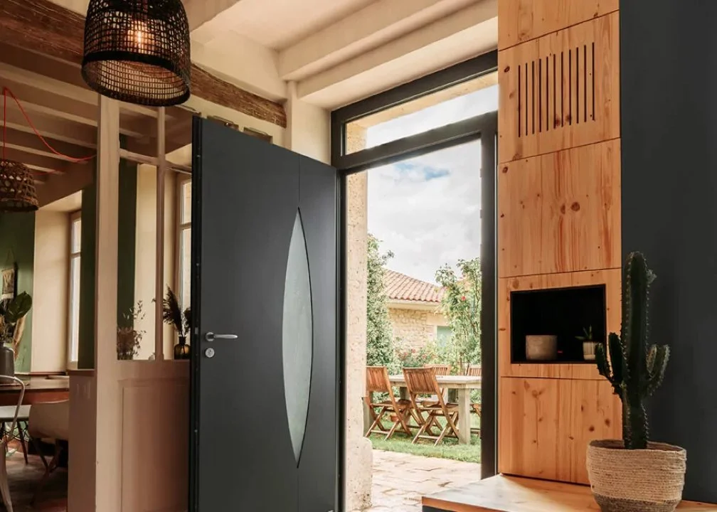 Une porte métallique au style moderne s'ouvre sur un patio extérieur avec une table et des chaises en bois, entouré de végétation et d'arbres, dans une maison avec un intérieur en bois et décorations naturelles.