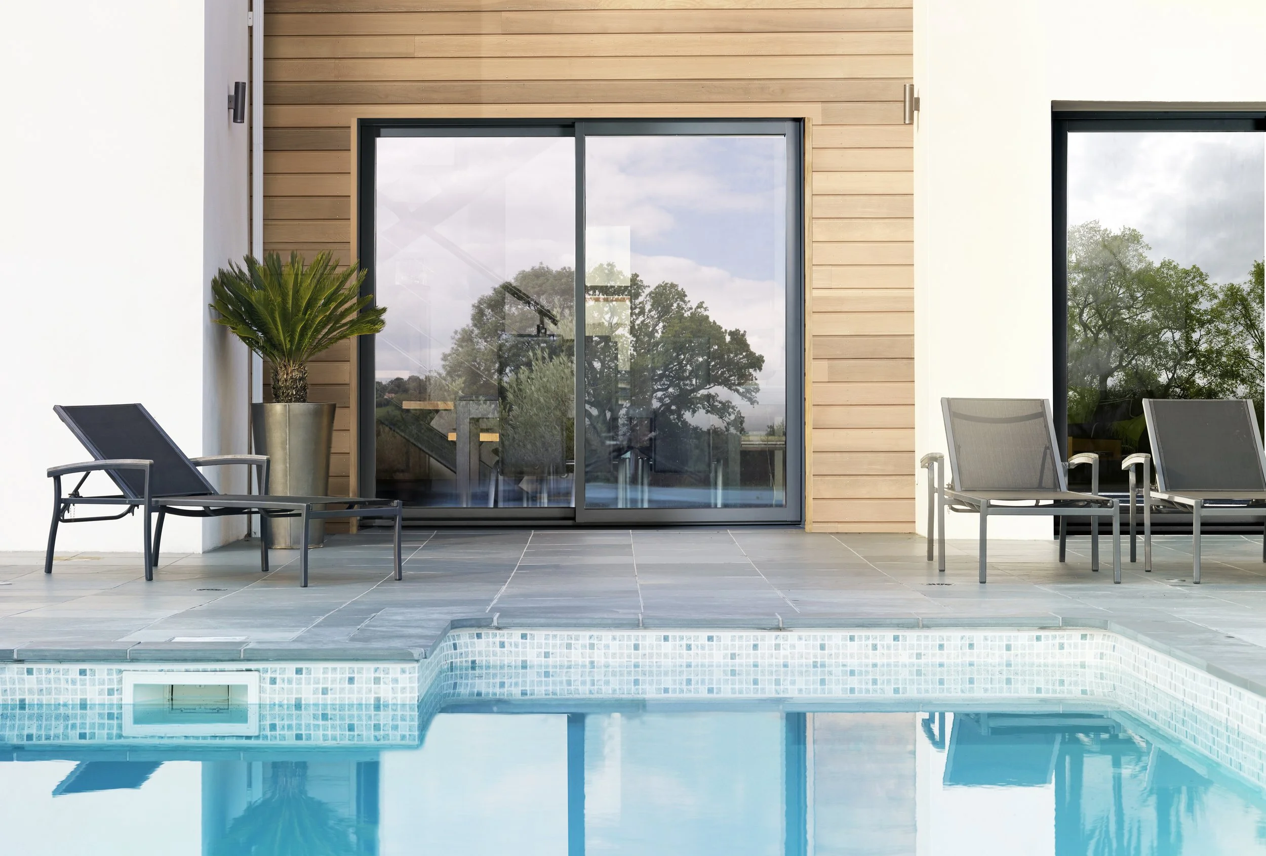 Terrasse avec chaises longues et piscine, façade de maison avec fenêtres en verre, bois et murs blancs, arbres visibles à travers les fenêtres.