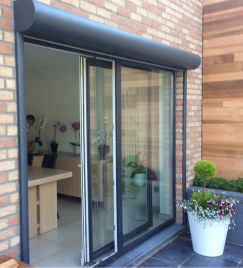 Une porte-fenêtre en verre avec une structure en aluminium, donnant sur un balcon ou une terrasse avec des plantes en pot, contre un mur en brique et en bois.