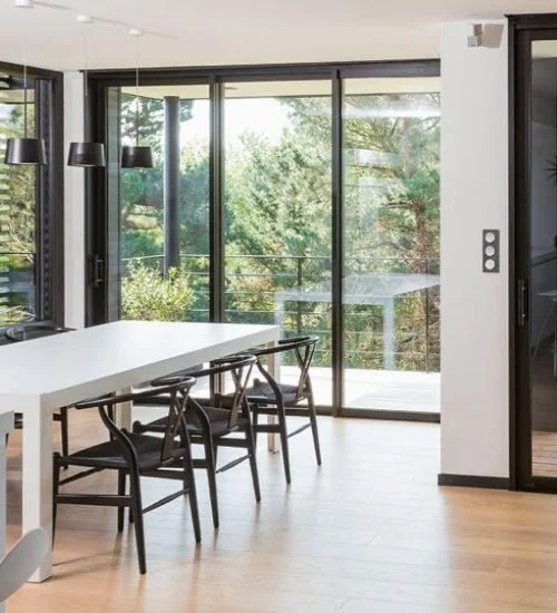 Salle à manger lumineuse avec une grande table blanche, six chaises en bois, et de grandes fenêtres donnant sur un balcon avec vue sur des arbres.