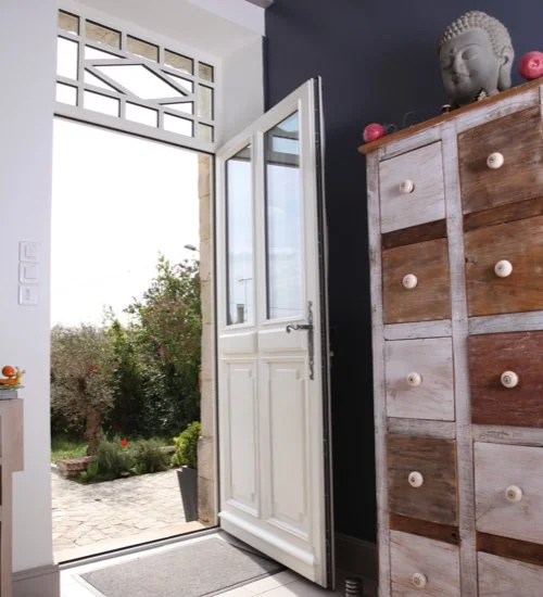 Une porte d'entrée blanche entrouverte donnant sur un jardin avec des buissons et des plantes ensoleillées, à côté d'une commode en bois avec un décor de tête de Bouddha.