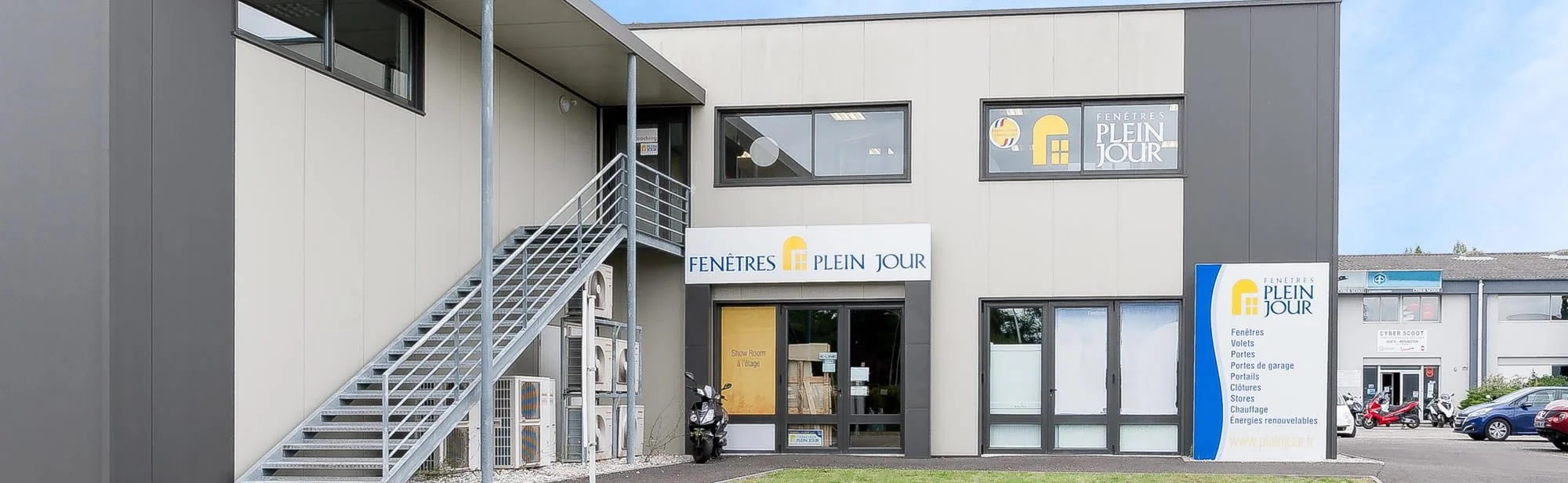 Façade d'un bâtiment commercial avec enseigne 'FENÊTRES PLEIN JOUR' et panneau indiquant les services offerts, comme fenêtres, volets, portes, garages, clôtures, chauffage, énergies renouvelables.