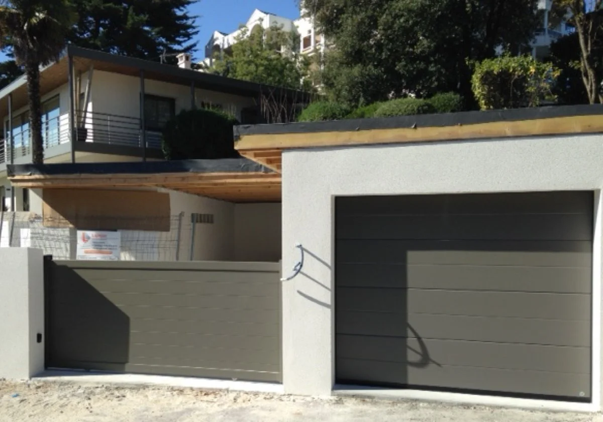 Une maison moderne avec un garage et une porte de garage en panneau gris, entourée d'un mur blanc et de végétation.