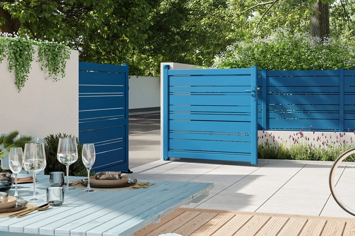 Terrasse extérieure avec table de pique-nique, verres, assiettes et couverts, clôture bleue, verdure et arbres en arrière-plan