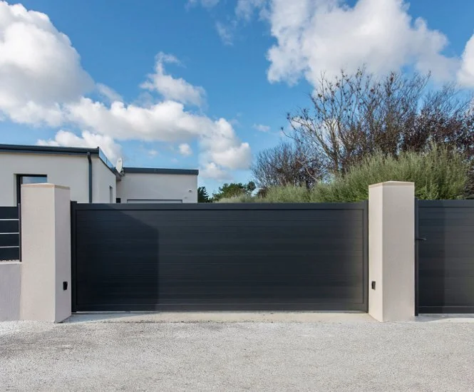 Portail noir en métal avec murs en béton, situé devant une maison moderne, avec un ciel bleu et des nuages.