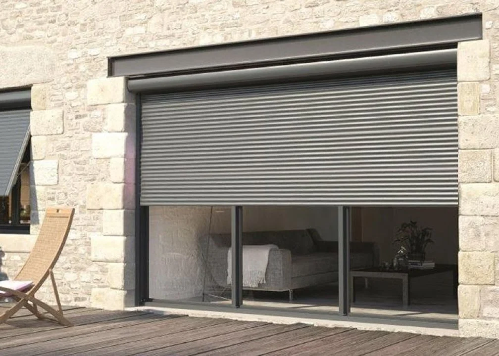 Fenêtre en partie fermée avec un volet roulant métallique sur une façade en pierre.