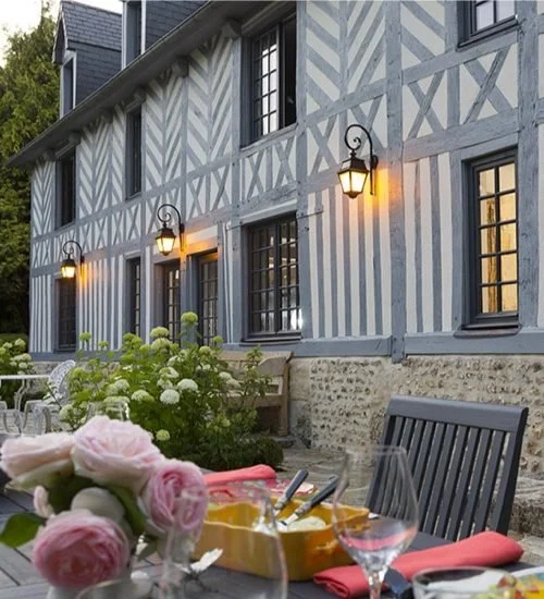 Terrasse avec fleurs, bouteille de vin et verre, en face d'une maison en colombage avec lumières allumées