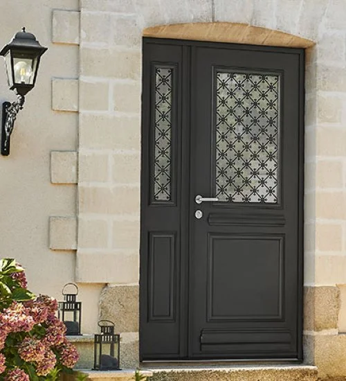 Porte d'entrée noire avec motifs en fer forgé, encadrée par un mur en pierre, avec une lampe murale à gauche et des fleurs en premier plan.